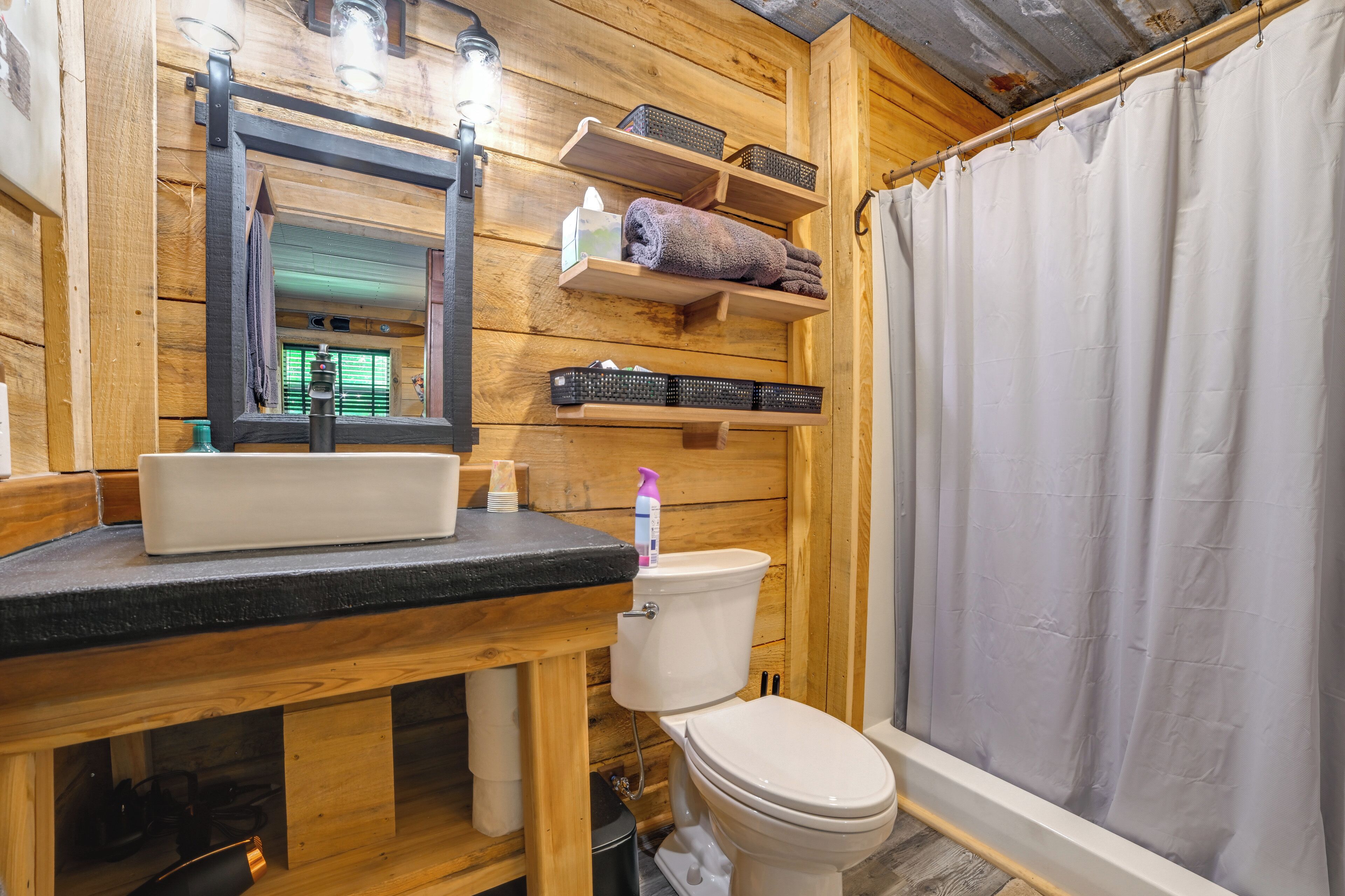 A wooden bathroom with a sink, mirror, shelves, and a toilet.