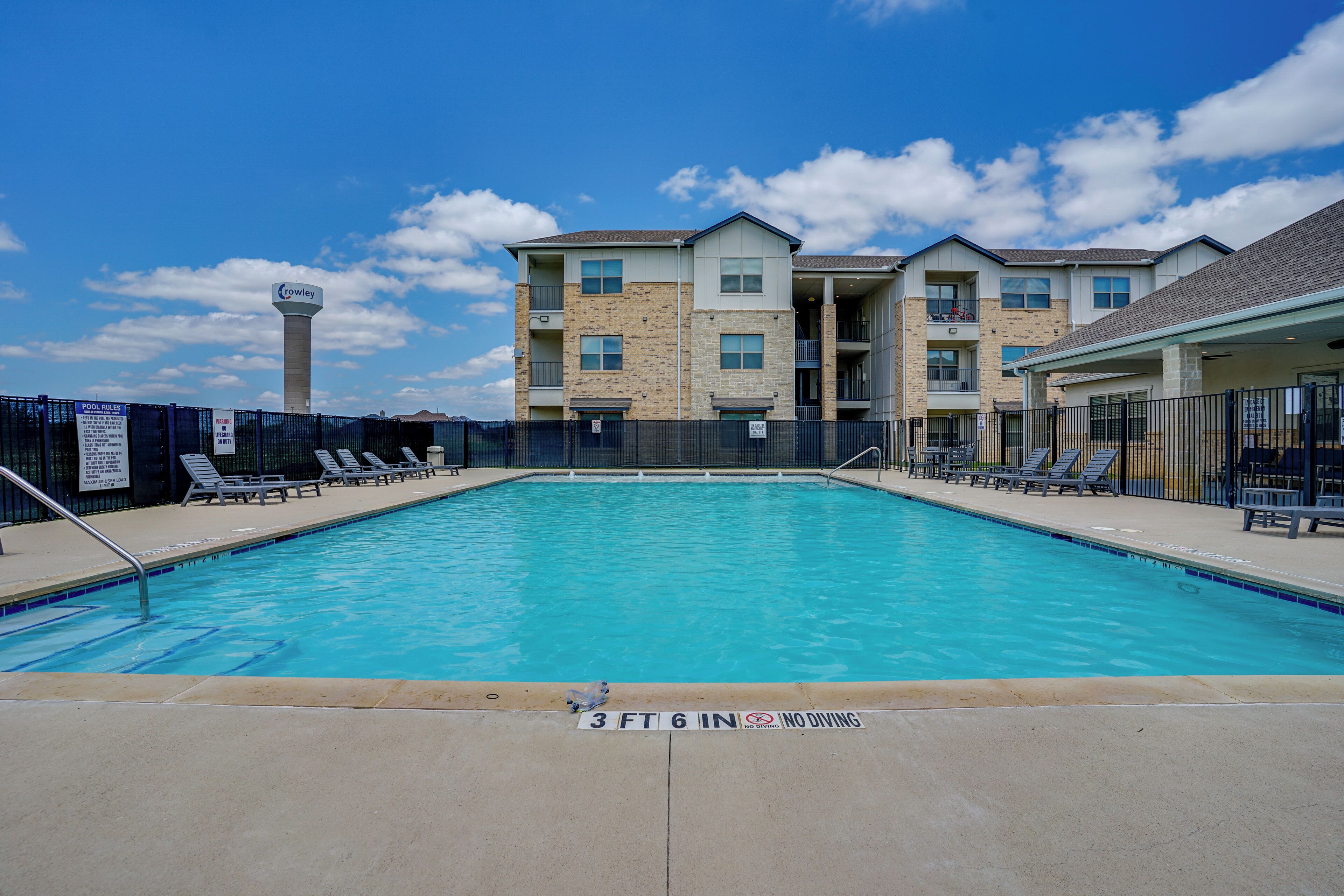 A swimming pool with lounge chairs, a sign indicating a depth of 3 ft 6 in, and a building with multiple balconies in the background.