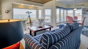 A living room with a sofa, a coffee table, and a lamp. There is a telescope on a tripod, a bookshelf, and a sign indicating a beach. - Direct Beach Access! Quiet Diamond Point Retreat (Sequim)