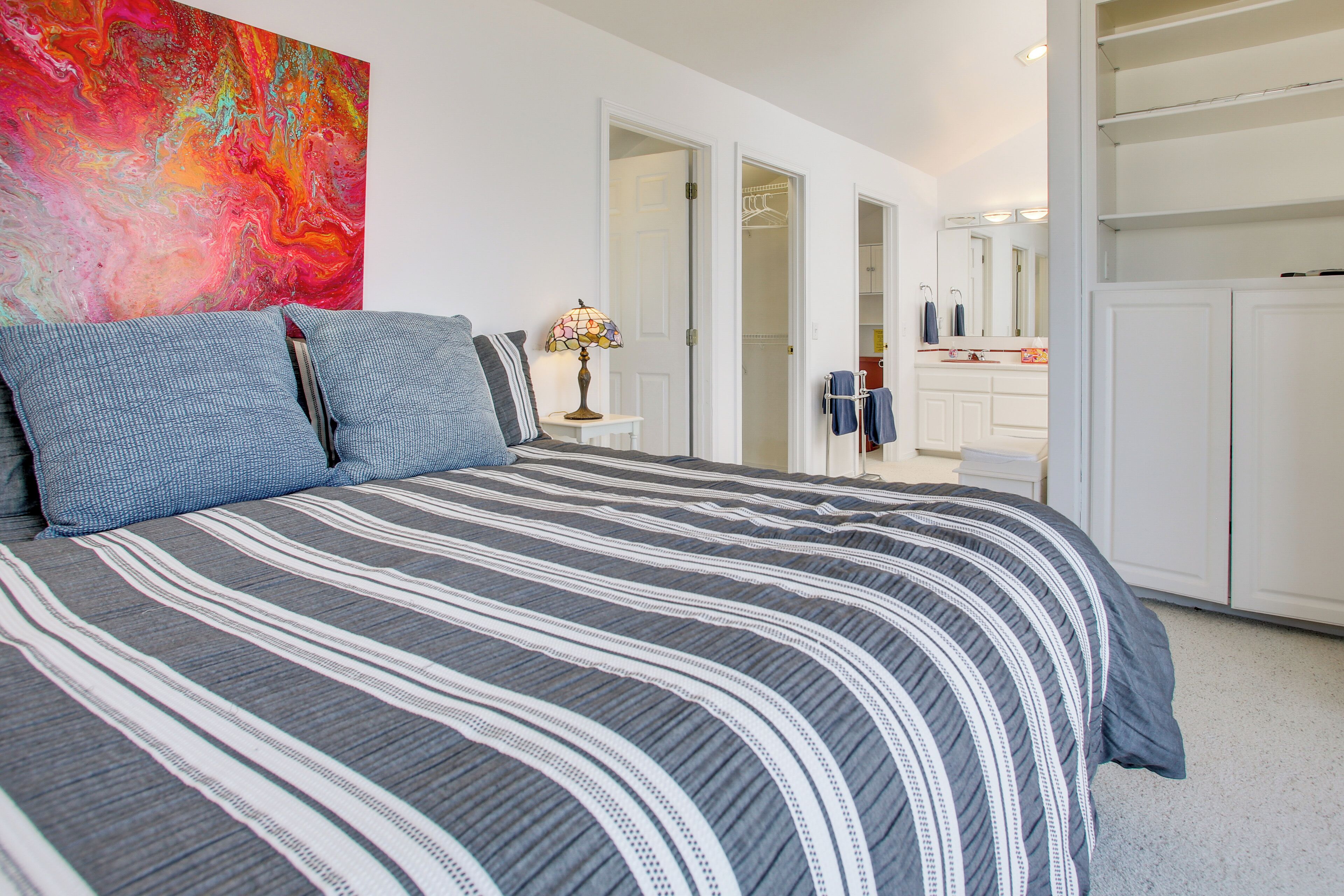 A bedroom with a bed, a colorful abstract painting, a bedside table with a lamp, and a closet with hanging clothes.