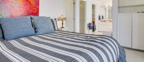A bedroom with a bed, a colorful abstract painting, a bedside table with a lamp, and a closet with hanging clothes.