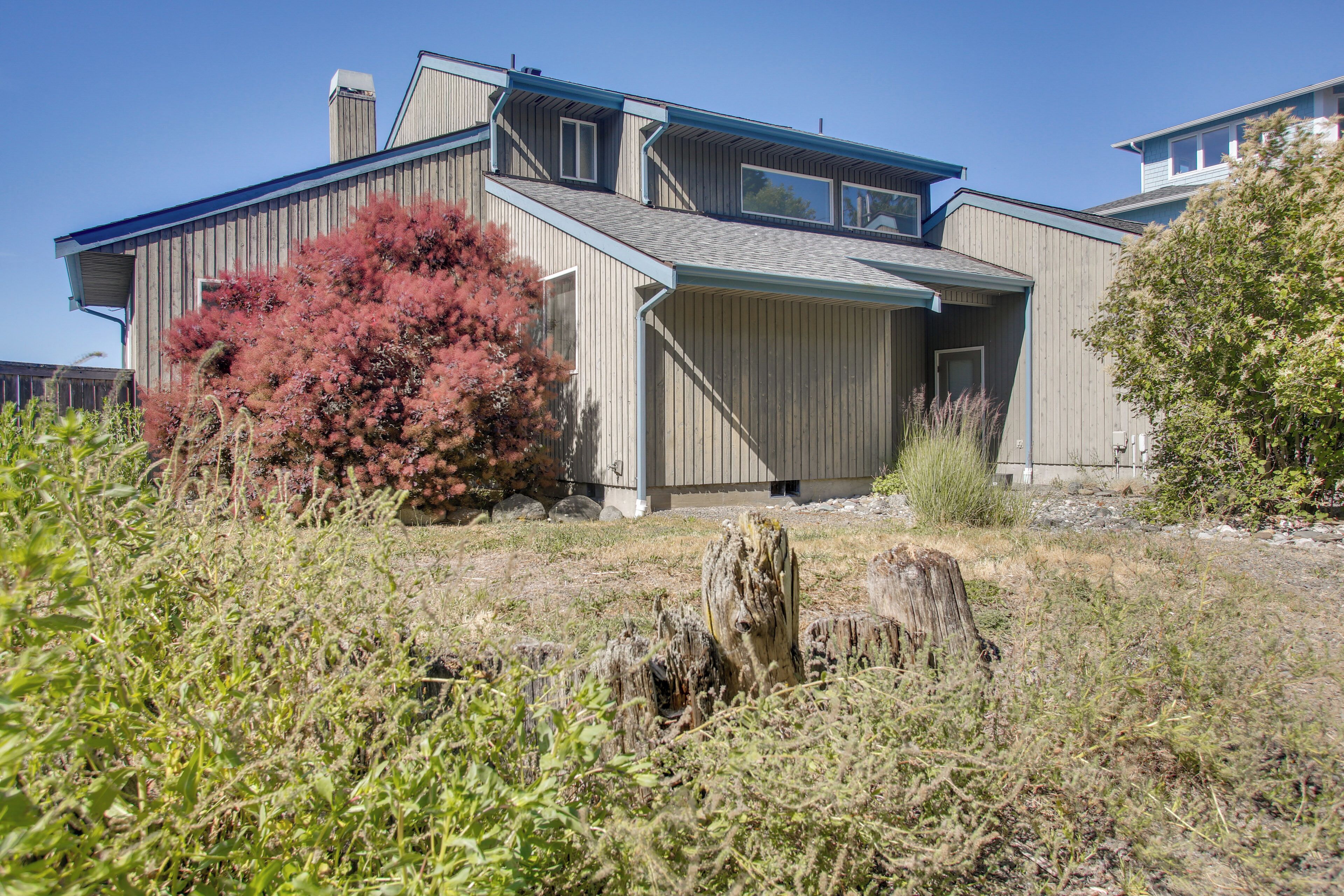 A modern two-story house with a large garage, a red flowering bush, and a prominent tree stump in the foreground.