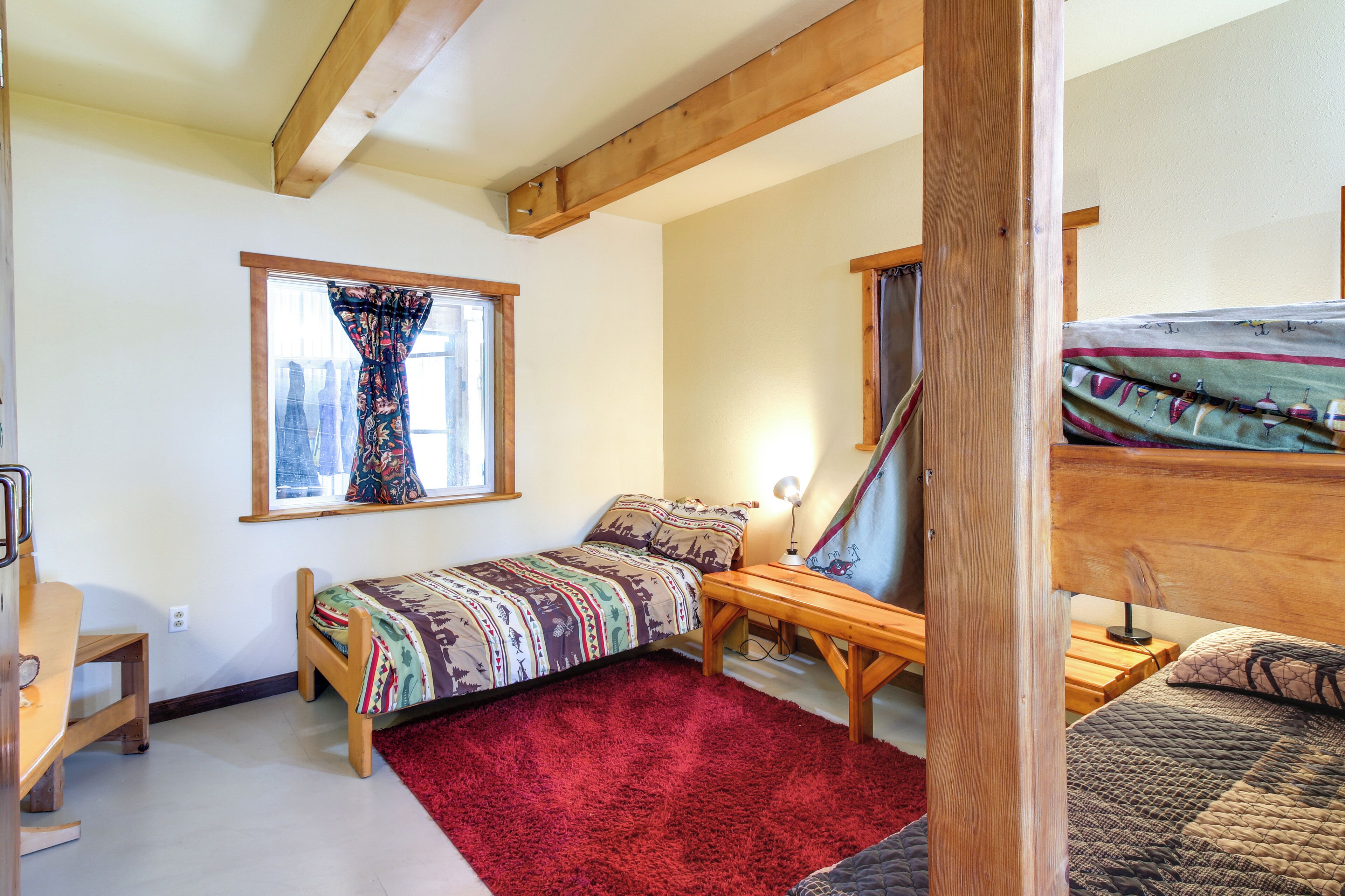 A bedroom with a single bed, a wooden desk, a red rug, and a window with curtains.