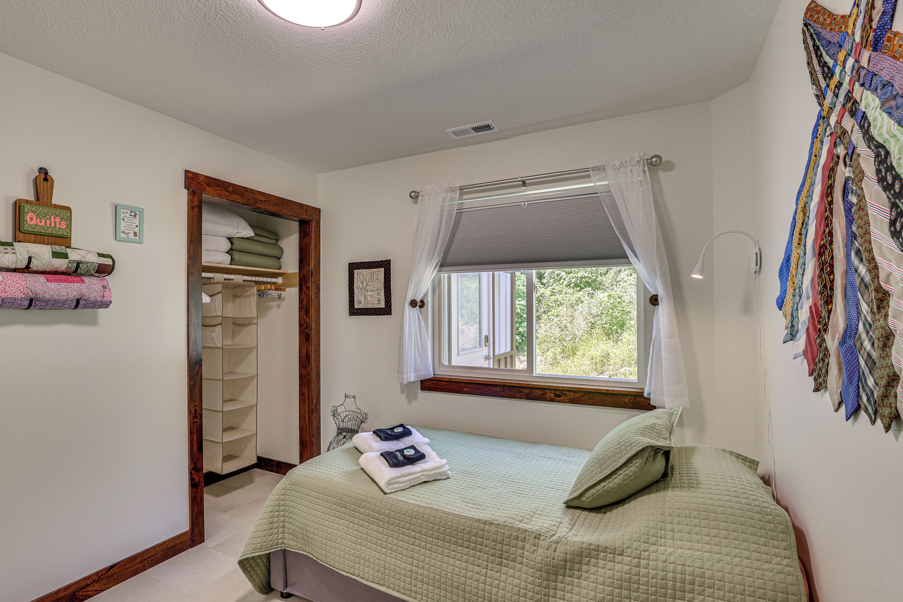 Un dormitorio con cama, ventana con cortinas, armario con ropa doblada y pared con colcha.