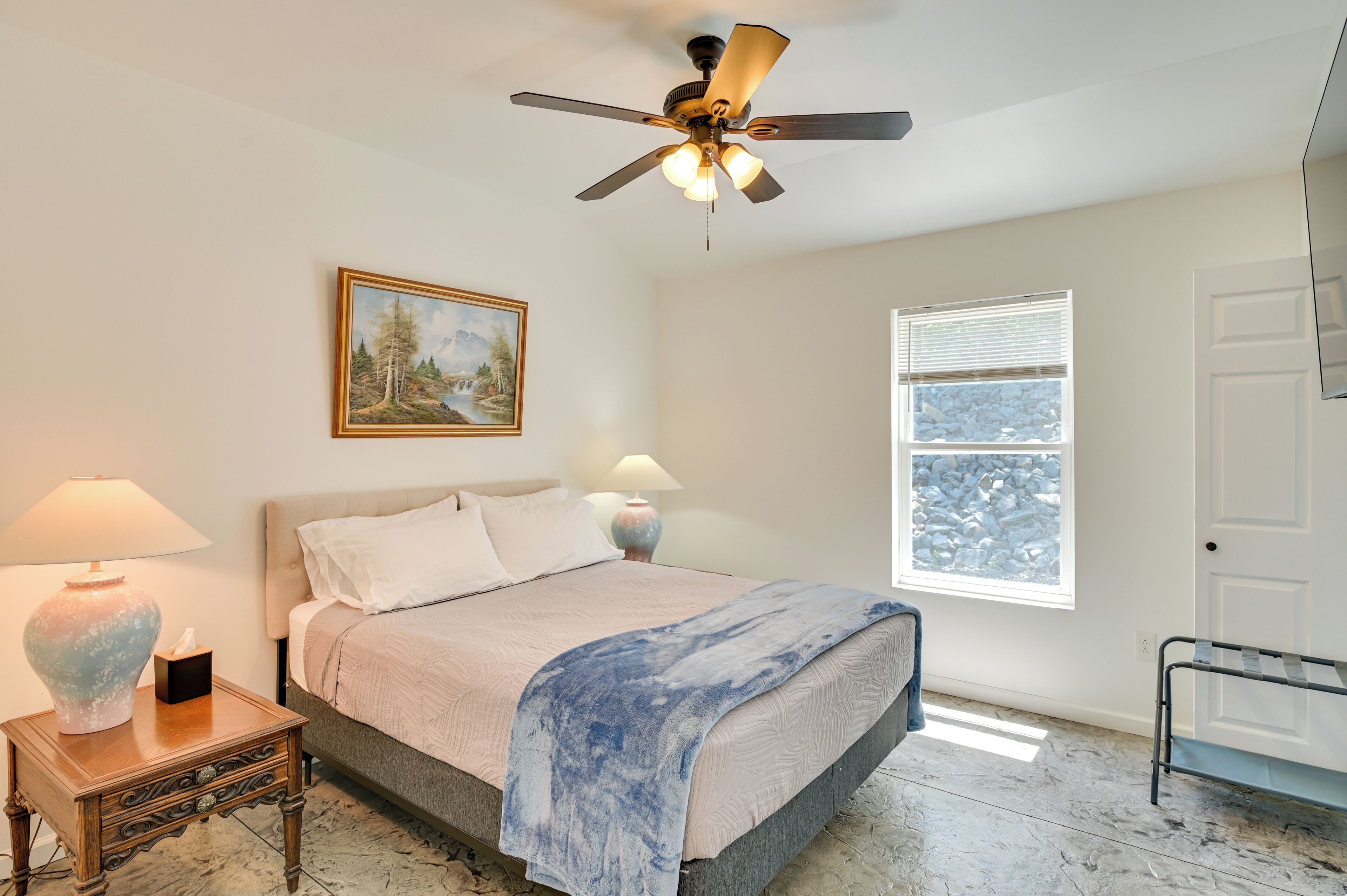 A bedroom with a bed, a ceiling fan, a window, a painting, and a nightstand with a lamp.