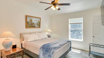 A bedroom with a bed, a ceiling fan, a window, a painting, and a nightstand with a lamp.