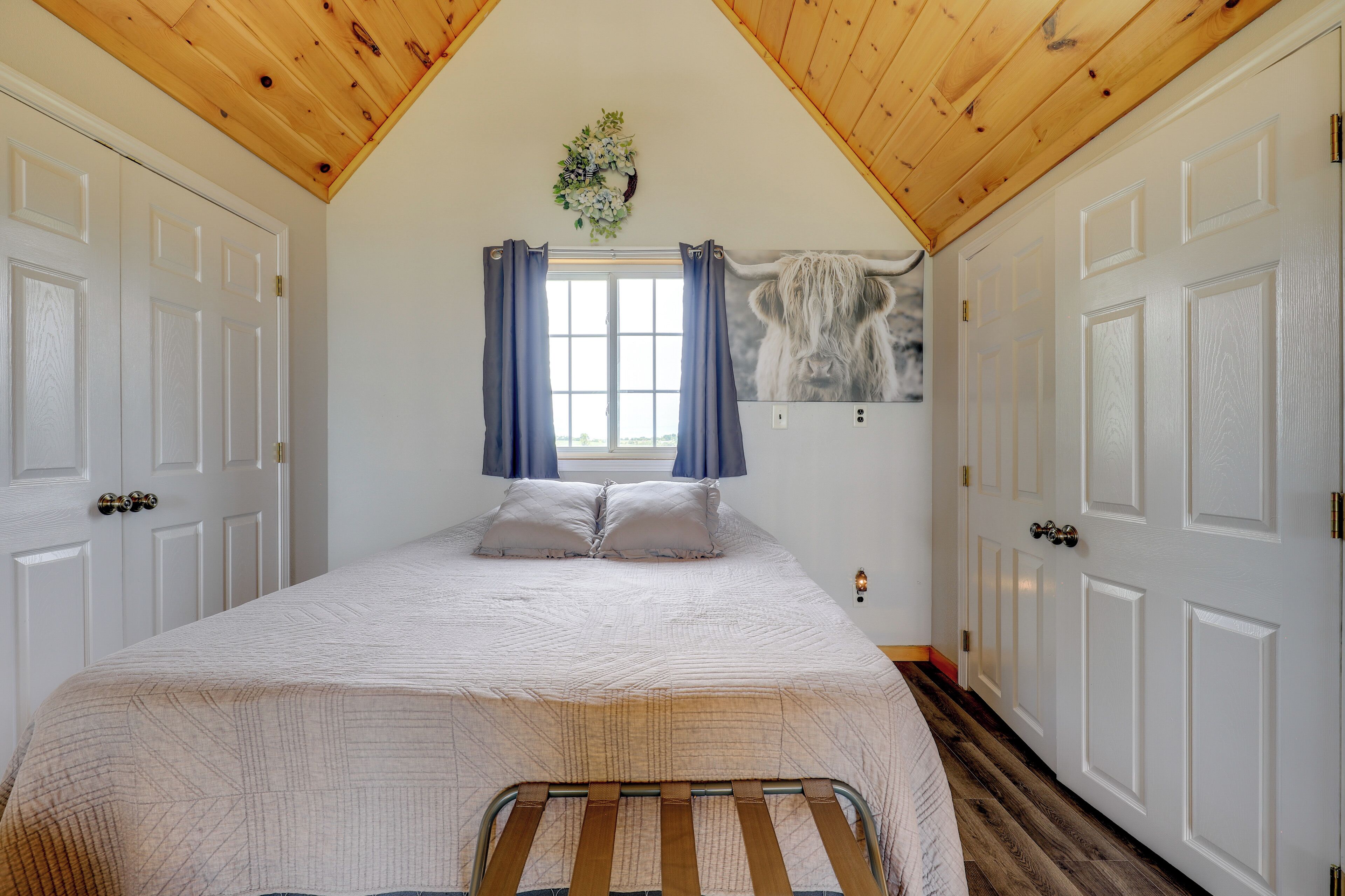 A bedroom with a bed, a window with curtains, a wall-mounted artwork, and wooden ceiling beams.