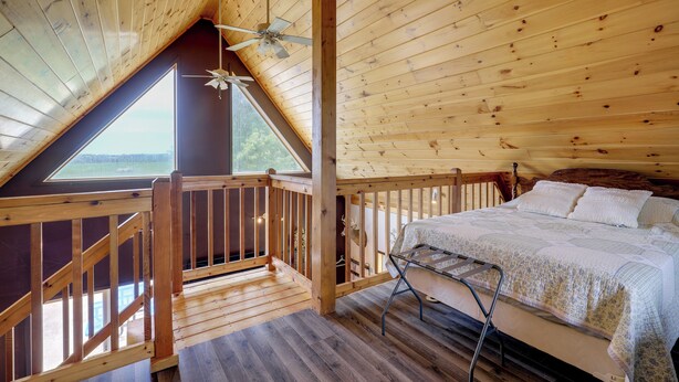 Un dormitorio con techo de madera, una cama con colcha, una mesita de noche y una ventana con vista a un paisaje.