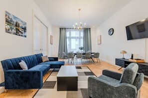 Ett modernt vardagsrum med en hörnsoffa, ett soffbord, en grå fåtölj, en matplats med bord och stolar, en kristallkrona och en platt-TV som är monterad på väggen.