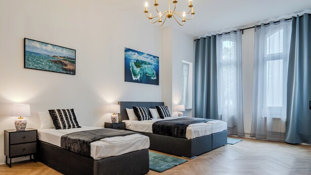 A modern bedroom with two beds, a chandelier, artwork on the walls, and a view of the outdoors. - Grand Living Apartment 150 m² 10 Guests 2 Bathrooms Ku'damm (Berlin)