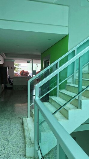 Eine Treppe mit Glasgeländern führt zu einer oberen Etage, der Boden ist mit Fliesen belegt und im Hintergrund ist eine grüne Wand zu sehen.