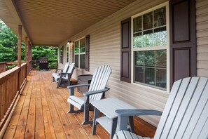 A covered porch with wooden flooring, white Adirondack chairs, and a grill.