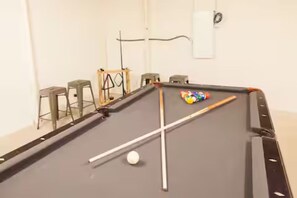 A pool table with a cue stick and balls, a small stool, and a wall-mounted device.