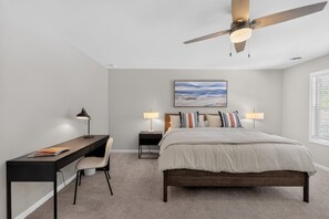 Ein Schlafzimmer mit Bett, Schreibtisch, Stuhl, zwei Nachttischen mit Lampen, einem Deckenventilator und einem Wandbild.