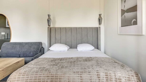 A bedroom with a bed, two white pillows, a grey headboard, a wooden bedside table, a grey sofa, and a wall-mounted picture. - Modern Studio 3P - Aubervilliers (Aubervilliers)