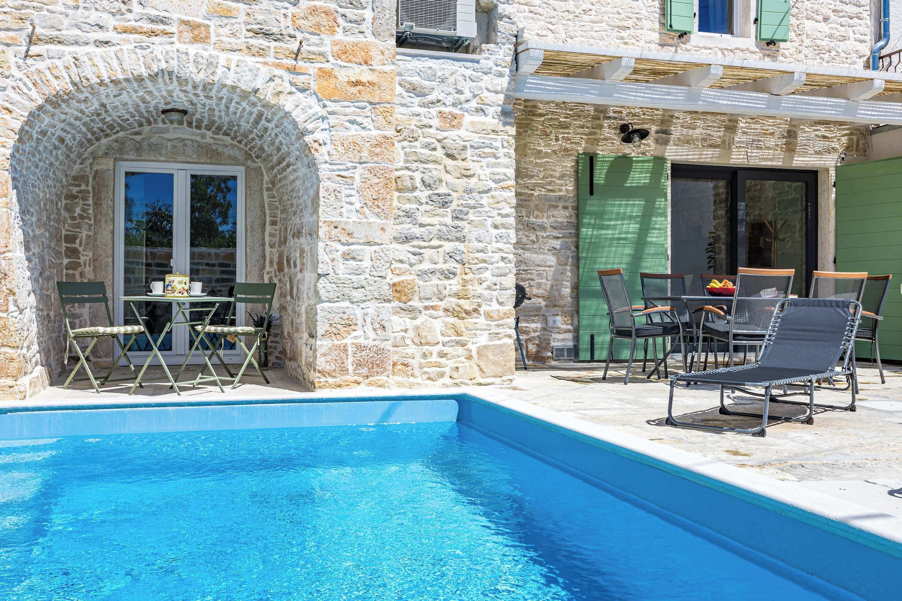 A stone building with a swimming pool, outdoor seating, and a door with a green shutter.
