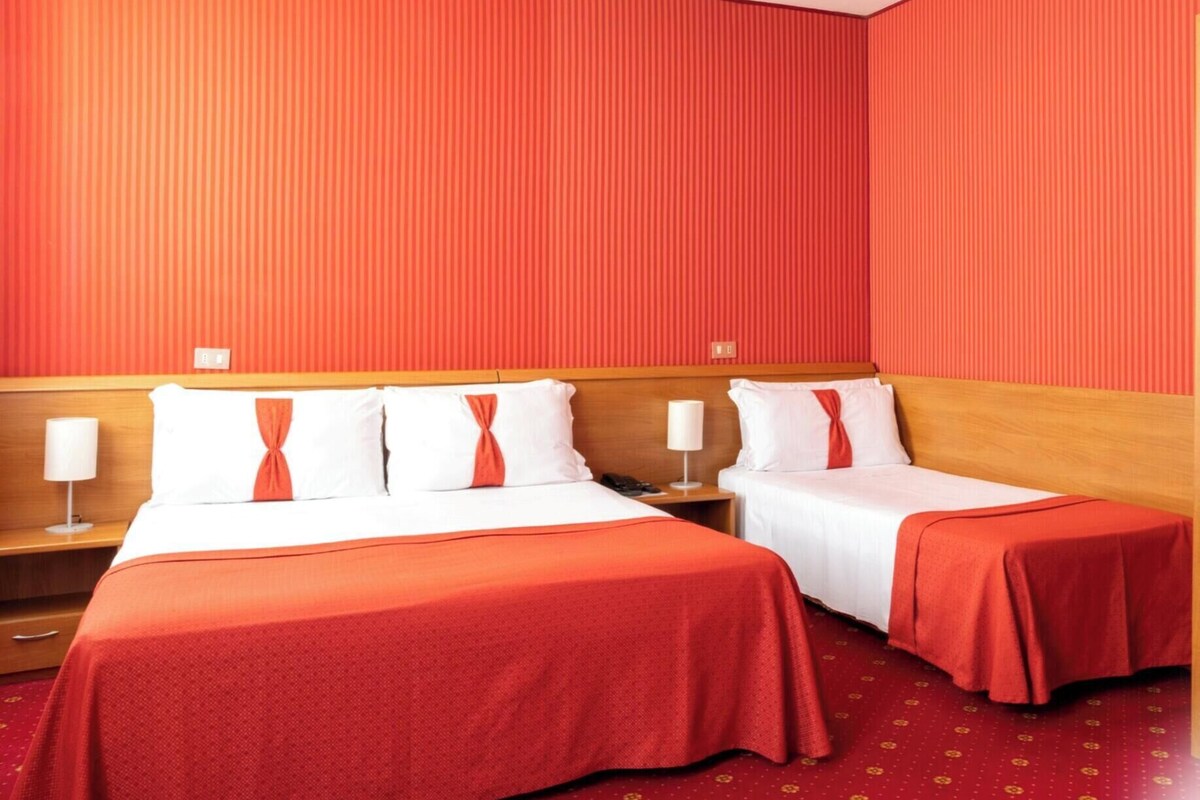 A hotel room with two beds, each with a red bedspread and white pillows. There are bedside tables with lamps, and the walls are covered with vertical red panels.
