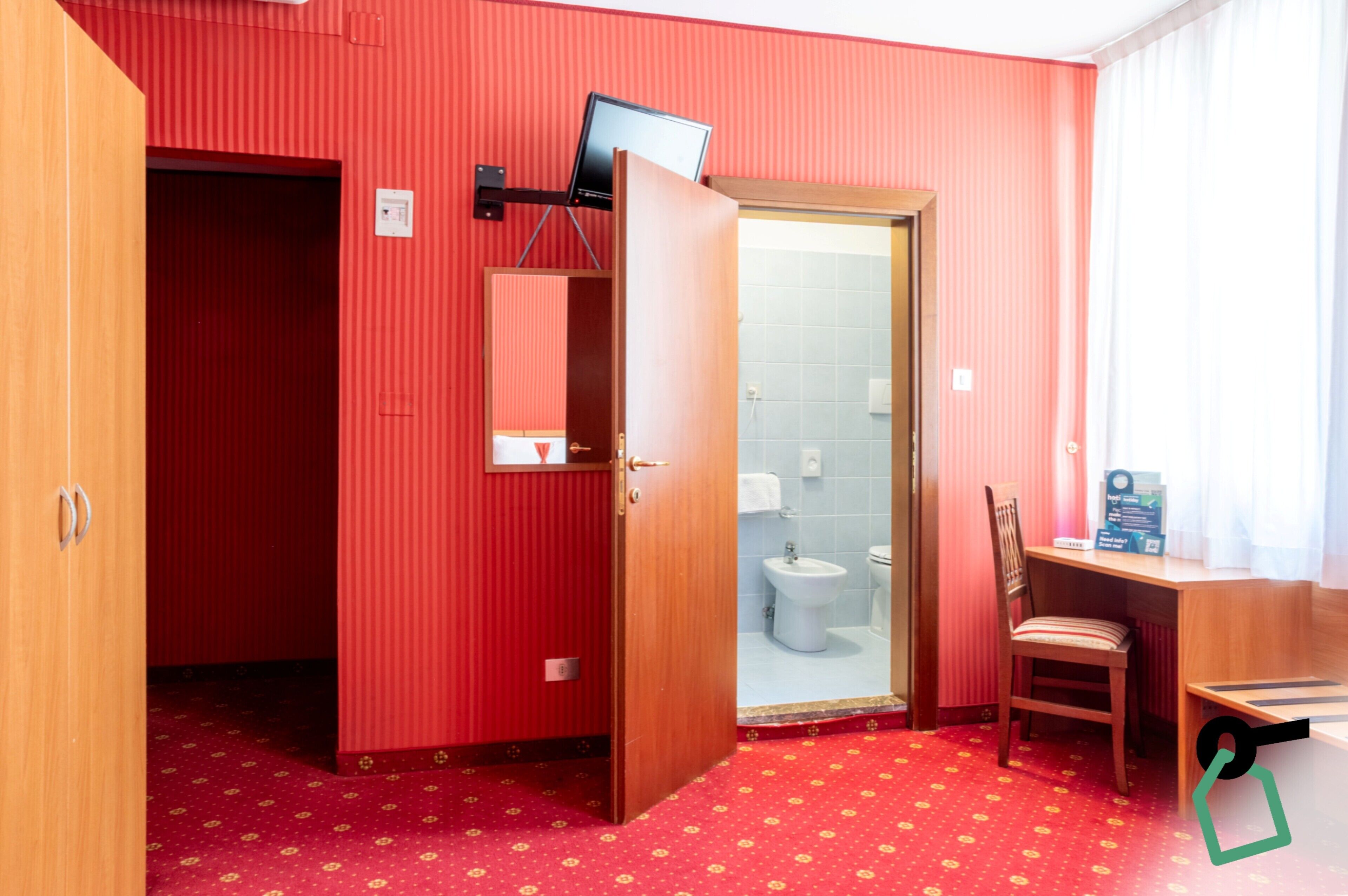 A compact room with a red patterned carpet, a wooden desk with a chair, a small bathroom with a toilet and bidet, and a mirror on the wall.