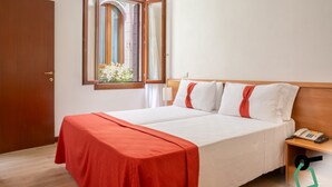 Cama doble con ropa de cama blanca y detalles rojos, cabecera de madera, mesita de noche con teléfono y ventana con cortinas.