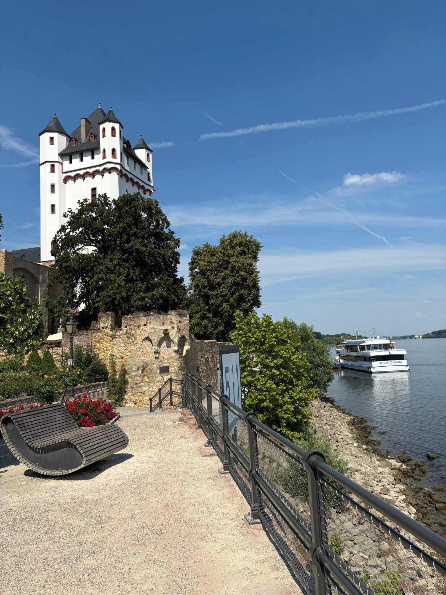 A castle with a tower, a boat on the water, and a paved walkway with a bench.