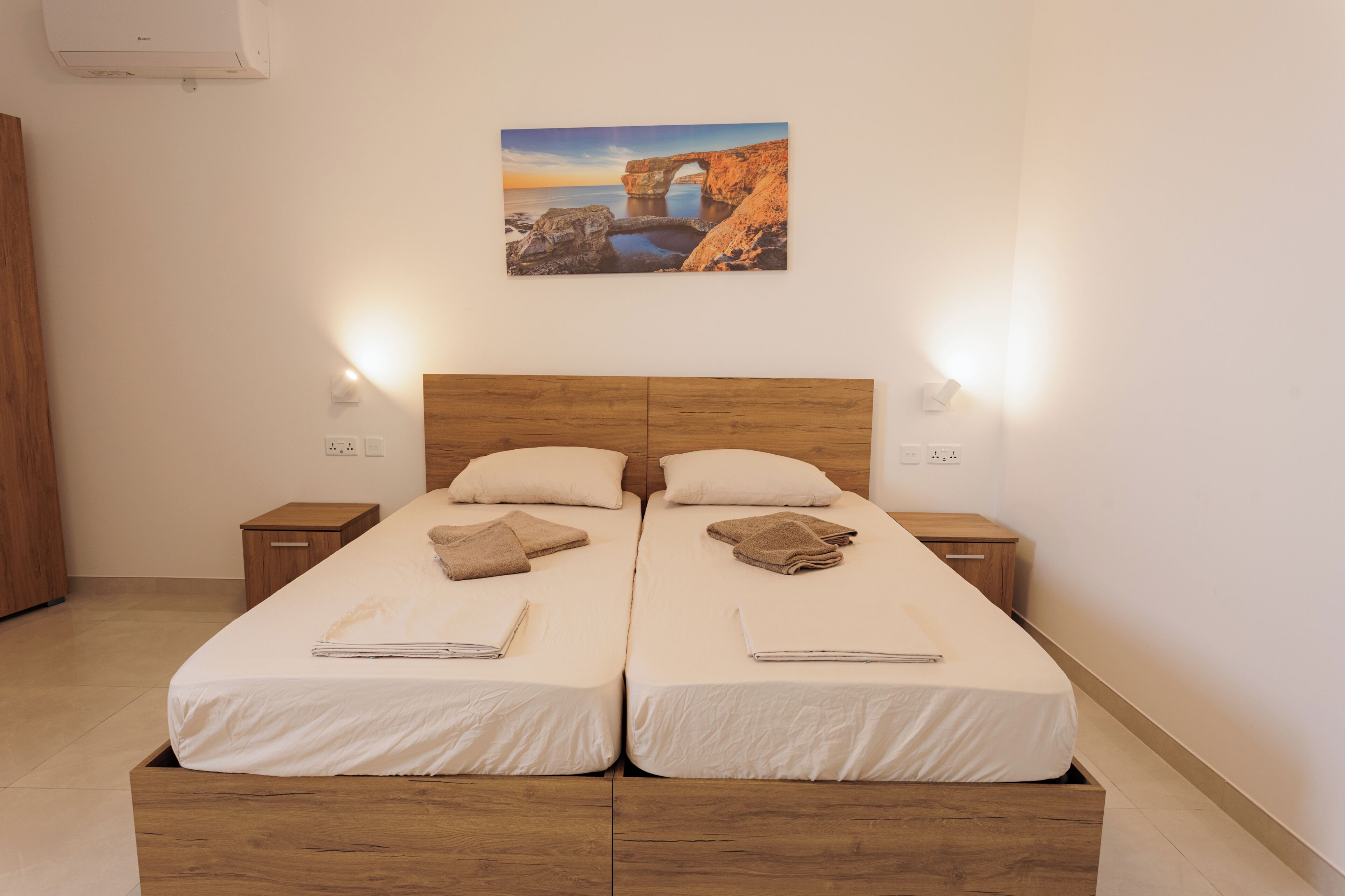 Een tweepersoonsbed met twee nachtkastjes, een houten hoofdbord en een schilderij van een kustlandschap.