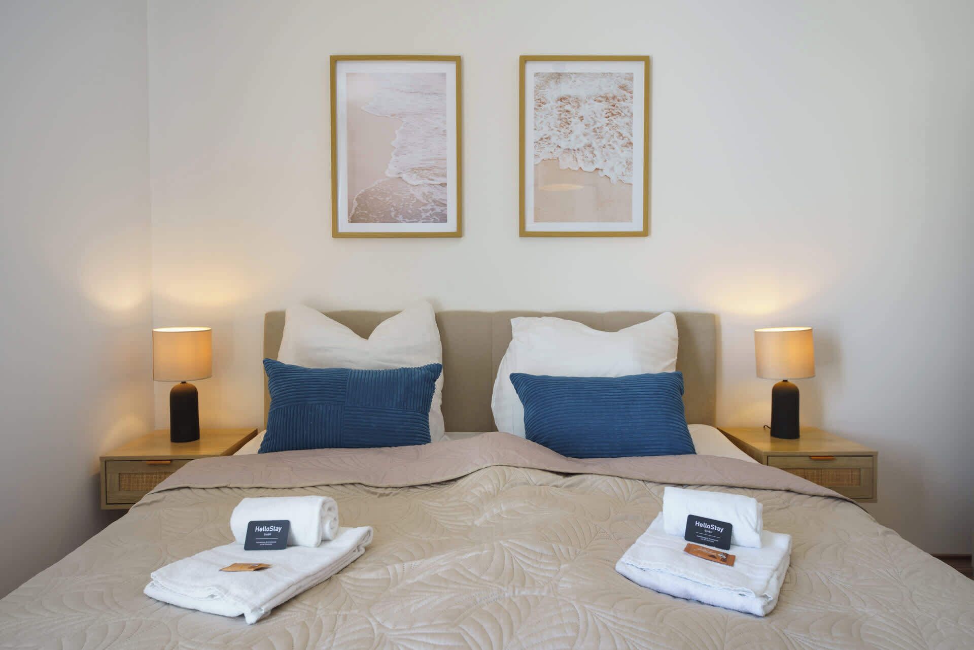 Ein Doppelbett mit zwei Kissen, einer Decke und zwei gefalteten Handtüchern mit dem 'HelloStay'-Etikett. Zwei Nachttischlampen und zwei gerahmte Bilder sind an der Wand zu sehen.