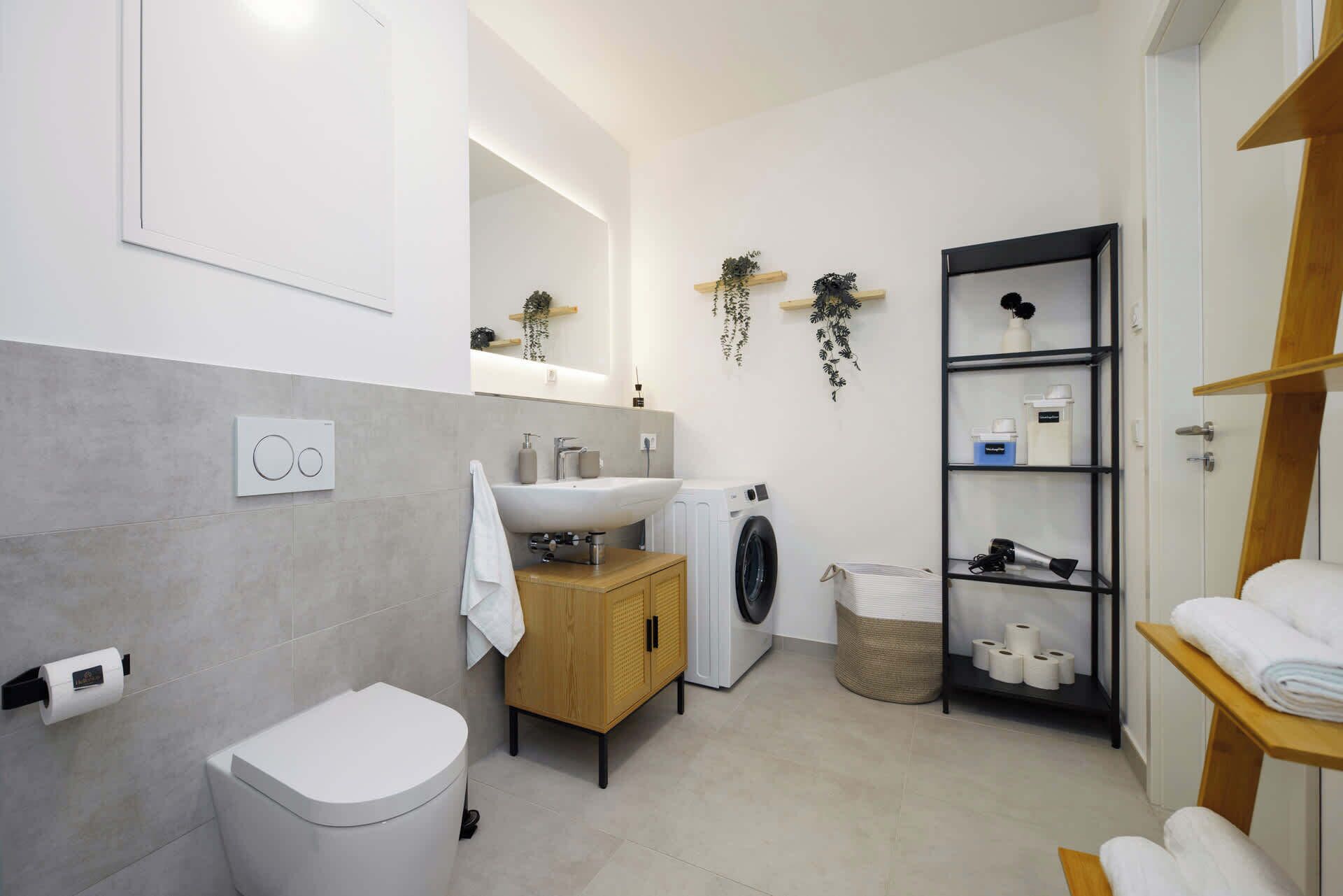 Ein modernes Badezimmer mit Toilette, Waschbecken, Waschmaschine und Regalen mit Pflegeprodukten.