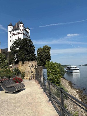 Un castillo con una torre, un barco en el agua y un paseo empedrado con un banco.