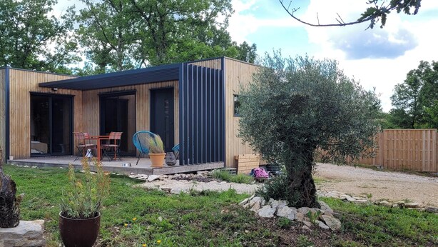 A modern wooden house with a patio, a dining area, and a garden with a tree and plants.