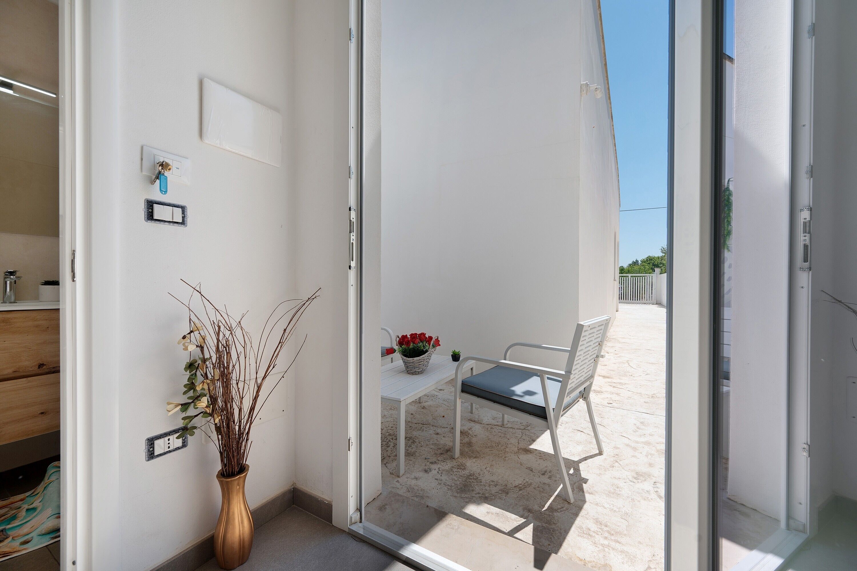 Una habitación con pared blanca, un armario de madera, un florero con ramas, una mesa y una silla, y vista a un balcón con cerca blanca.