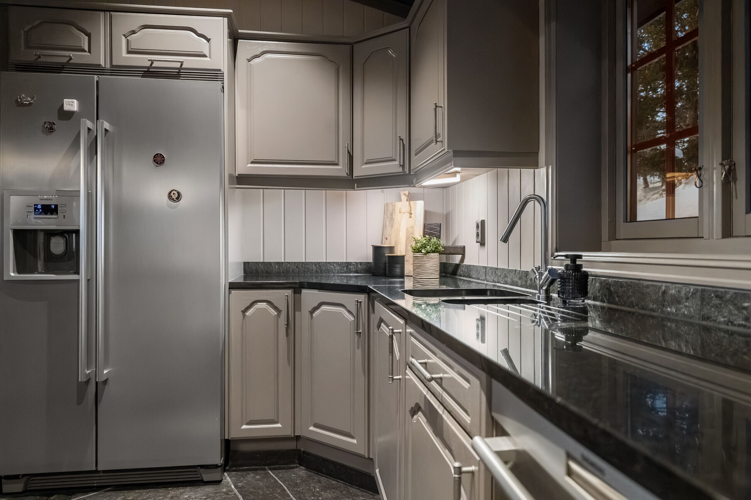 A kitchen with white cabinetry, a stainless steel refrigerator, a granite countertop, and a window with a view of trees.