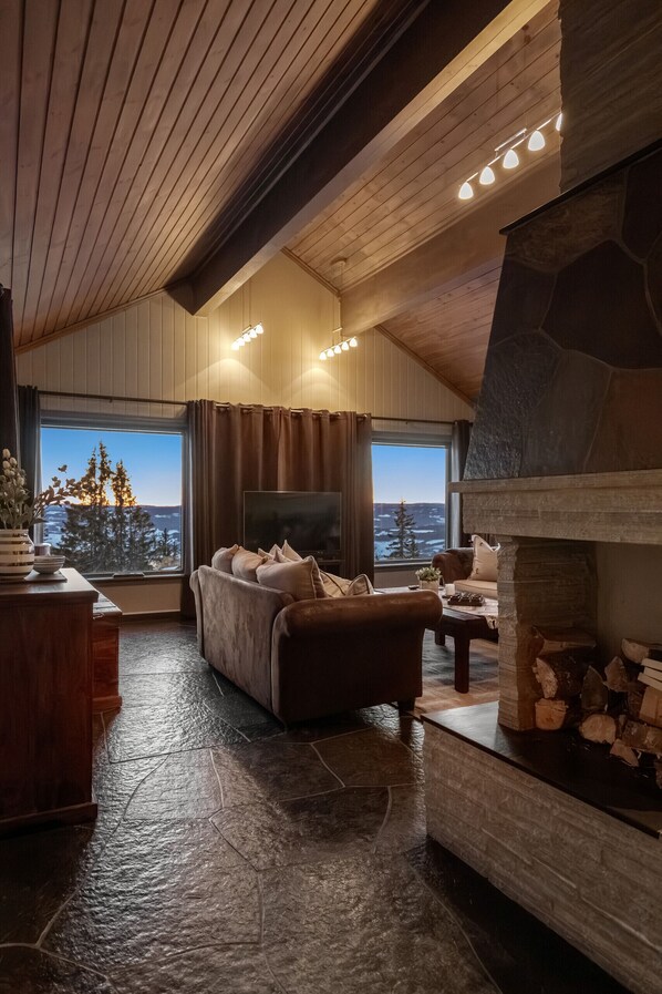 A cozy living room with a stone fireplace, a large window with a view of trees and hills, and a comfortable seating area.