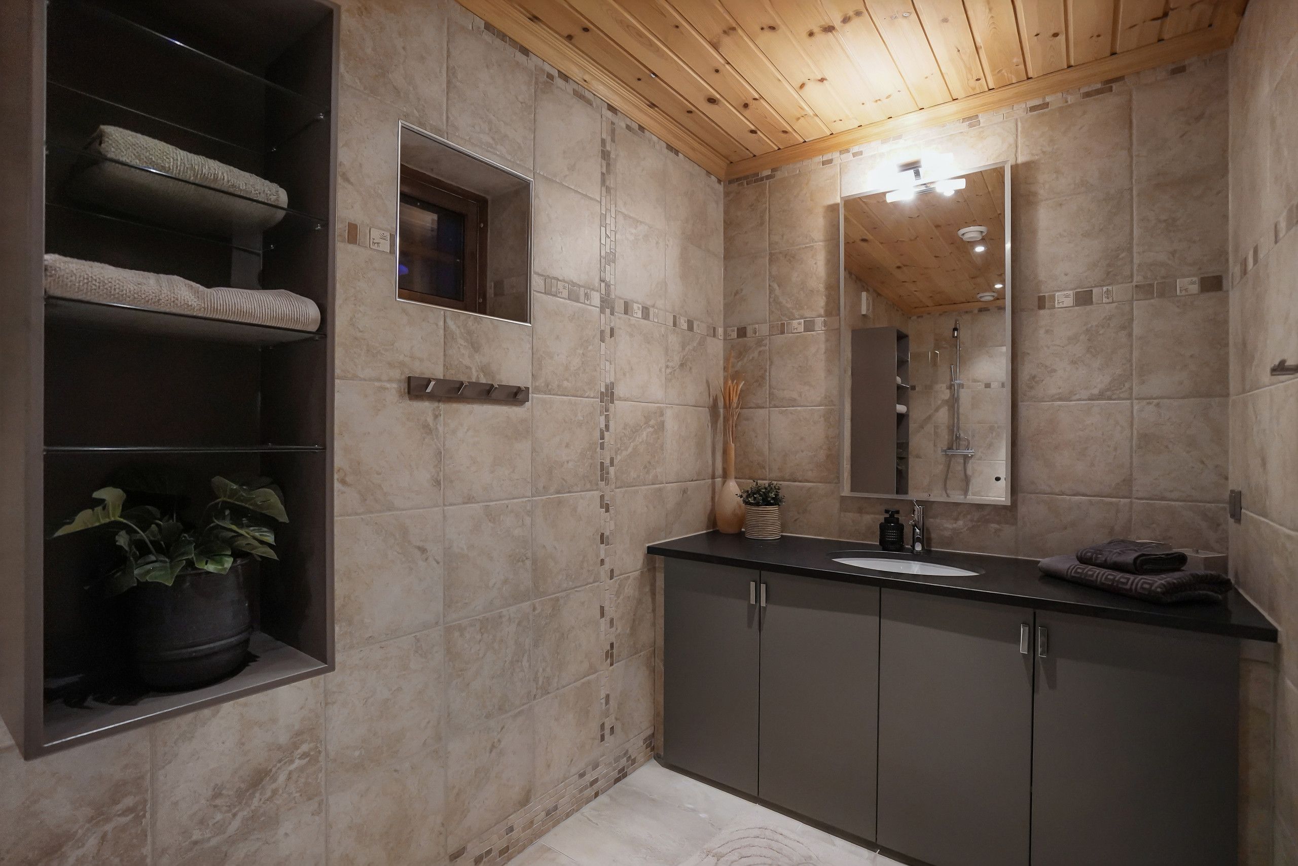 A bathroom with a wooden ceiling, a sink, a mirror, and a shelf with towels.