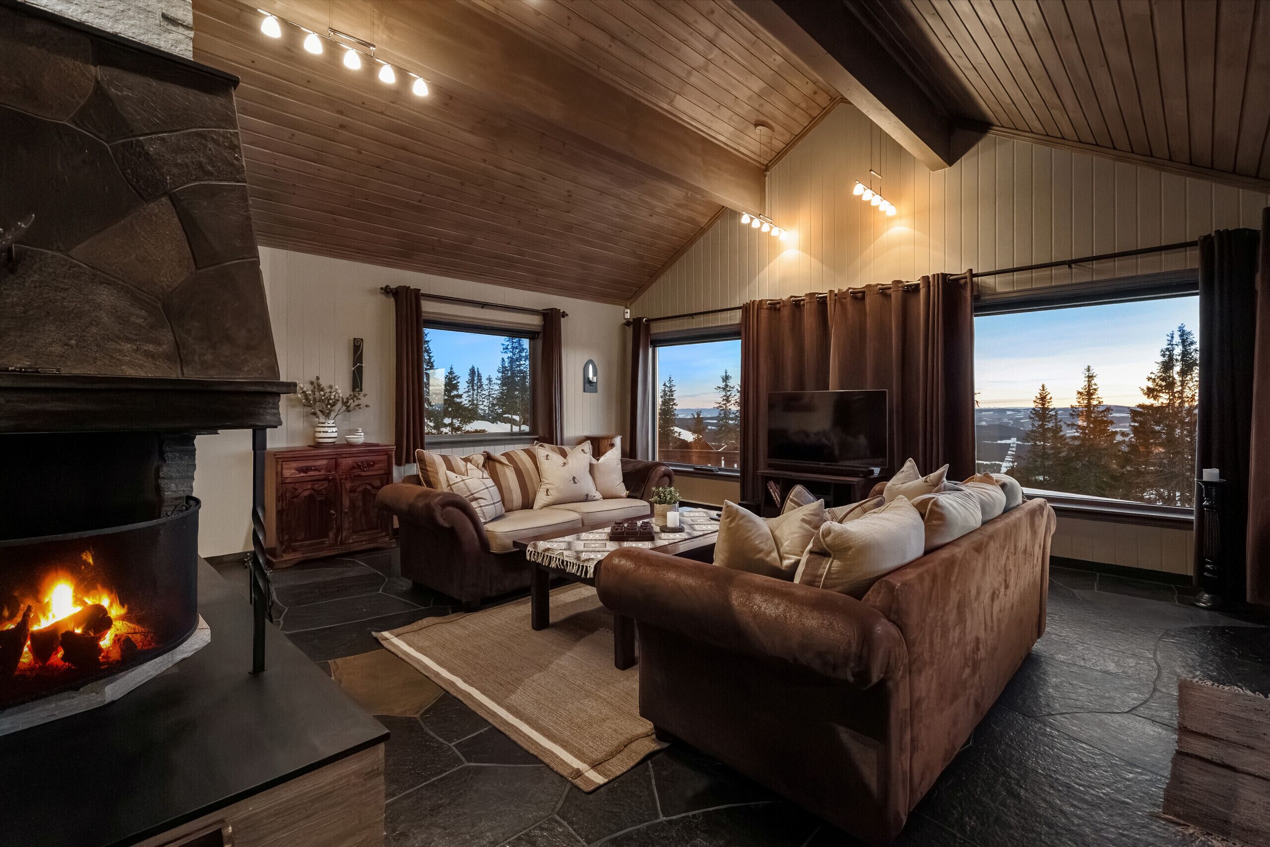 A living room with a stone fireplace, leather sofa, coffee table, and large windows with a view of trees.