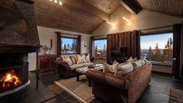 A living room with a stone fireplace, leather sofa, coffee table, and large windows with a view of trees.