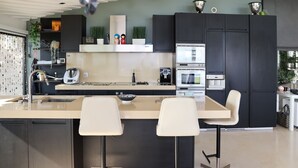 Et moderne køkken med en central ø, to barstole, indbygget ovn og kaffemaskine.