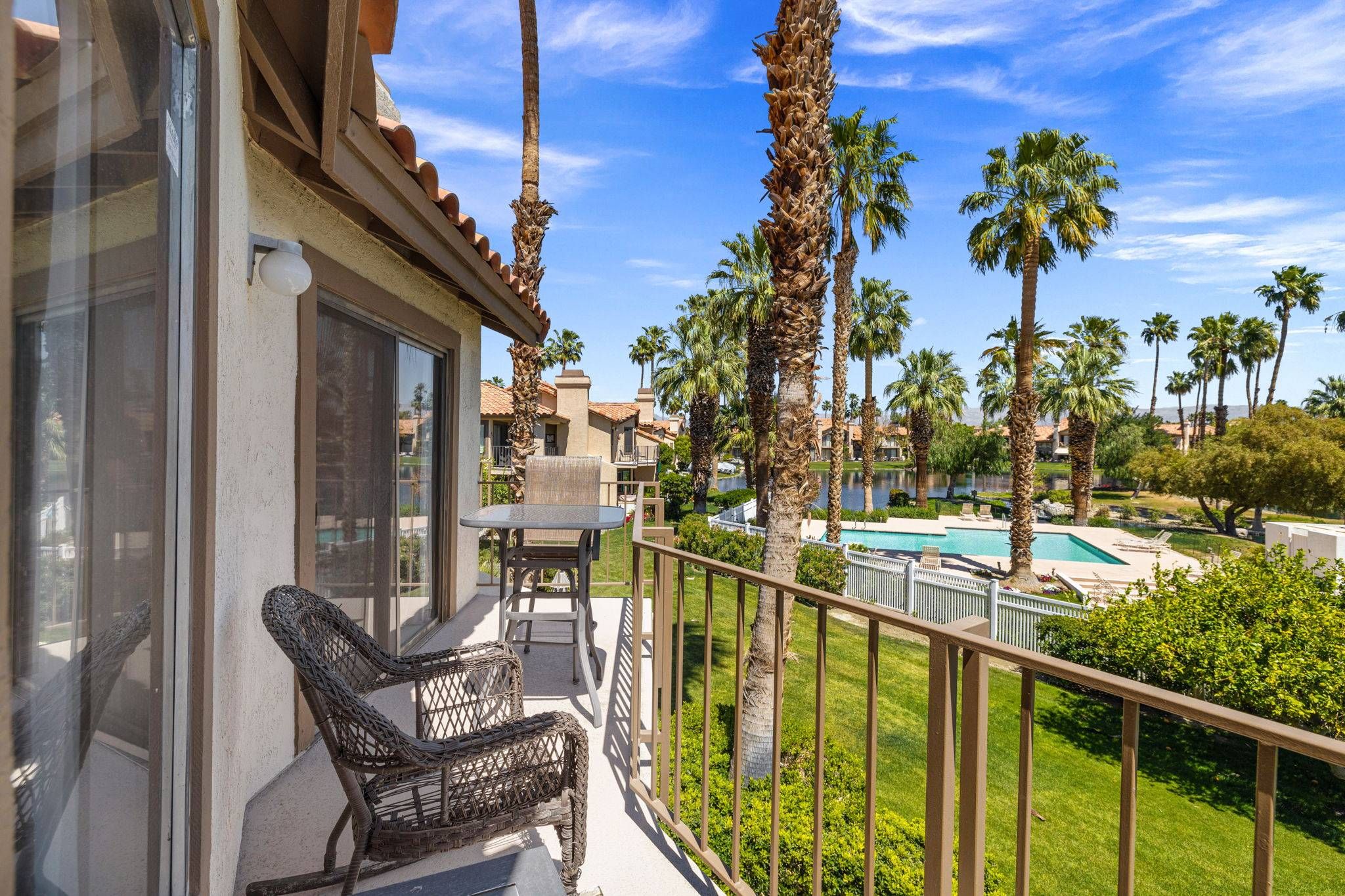 A balcony with a wicker chair and a table, overlooking a pool and palm trees.
