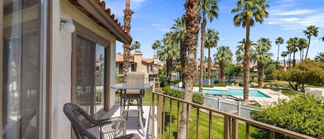 A balcony with a wicker chair and a table, overlooking a pool and palm trees.