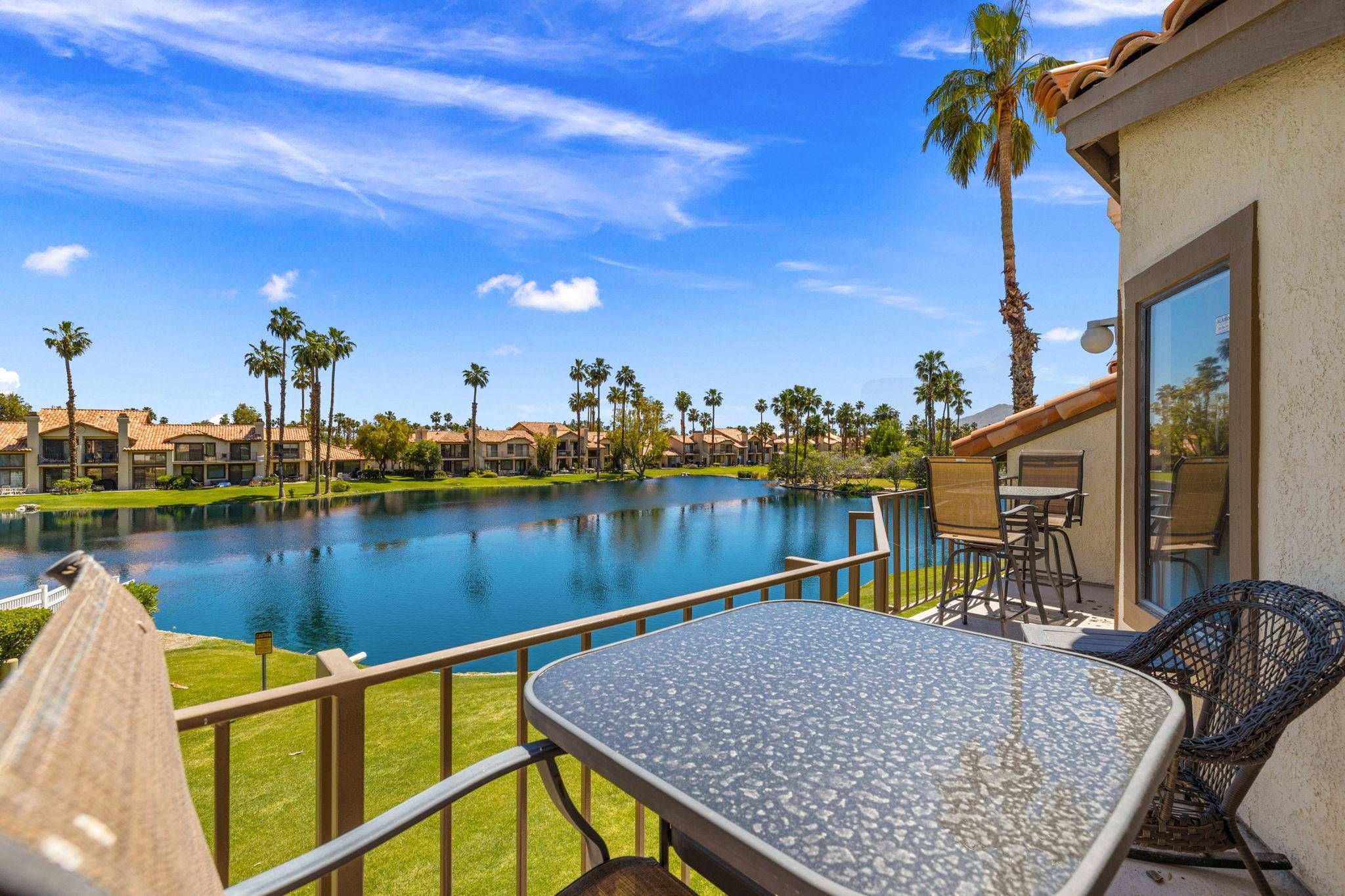 A poolside area with a clear view of a swimming pool, surrounded by palm trees and a building with a tiled roof.