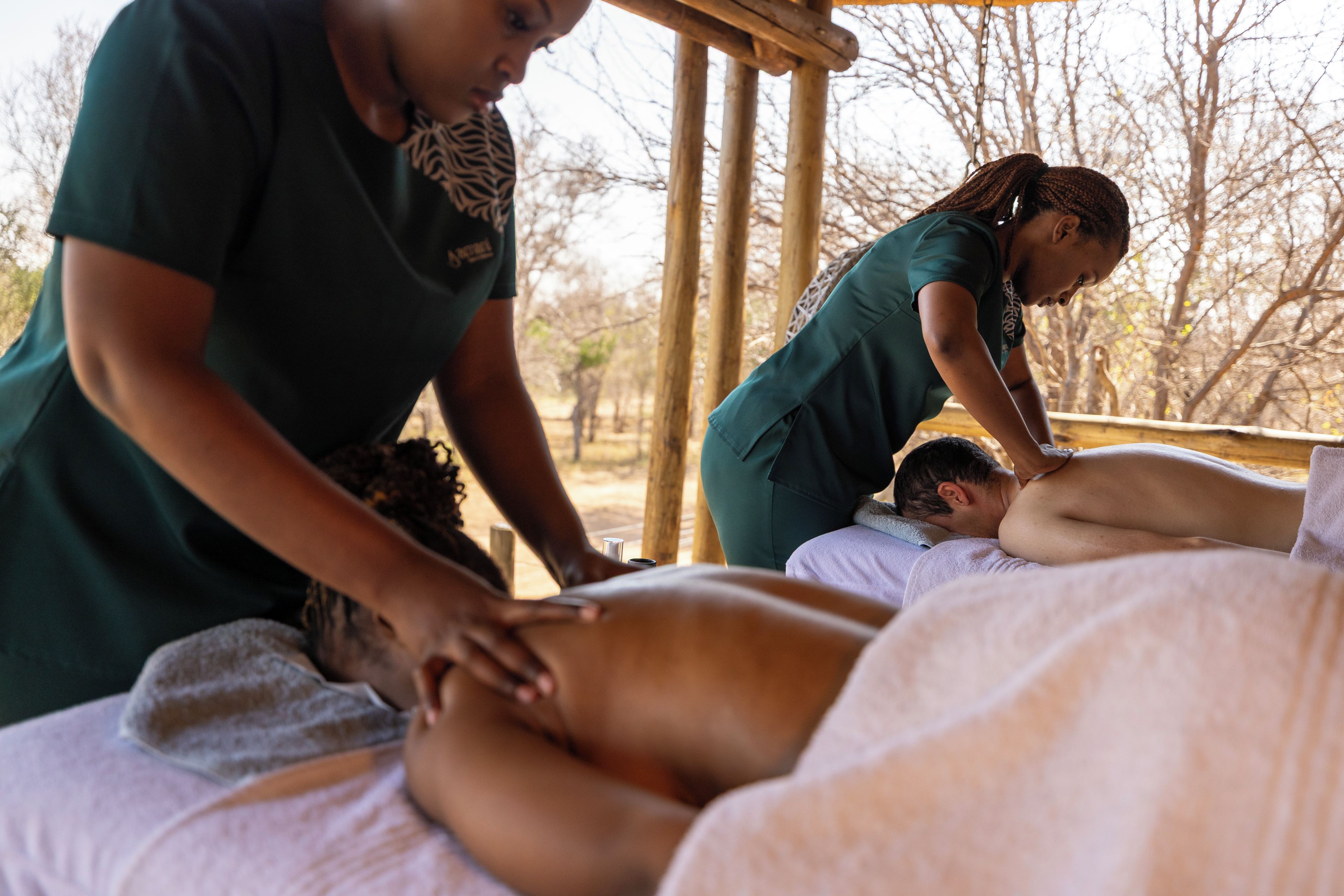 En massagebehandling i en naturnära miljö med träkonstruktioner och träd i bakgrunden.