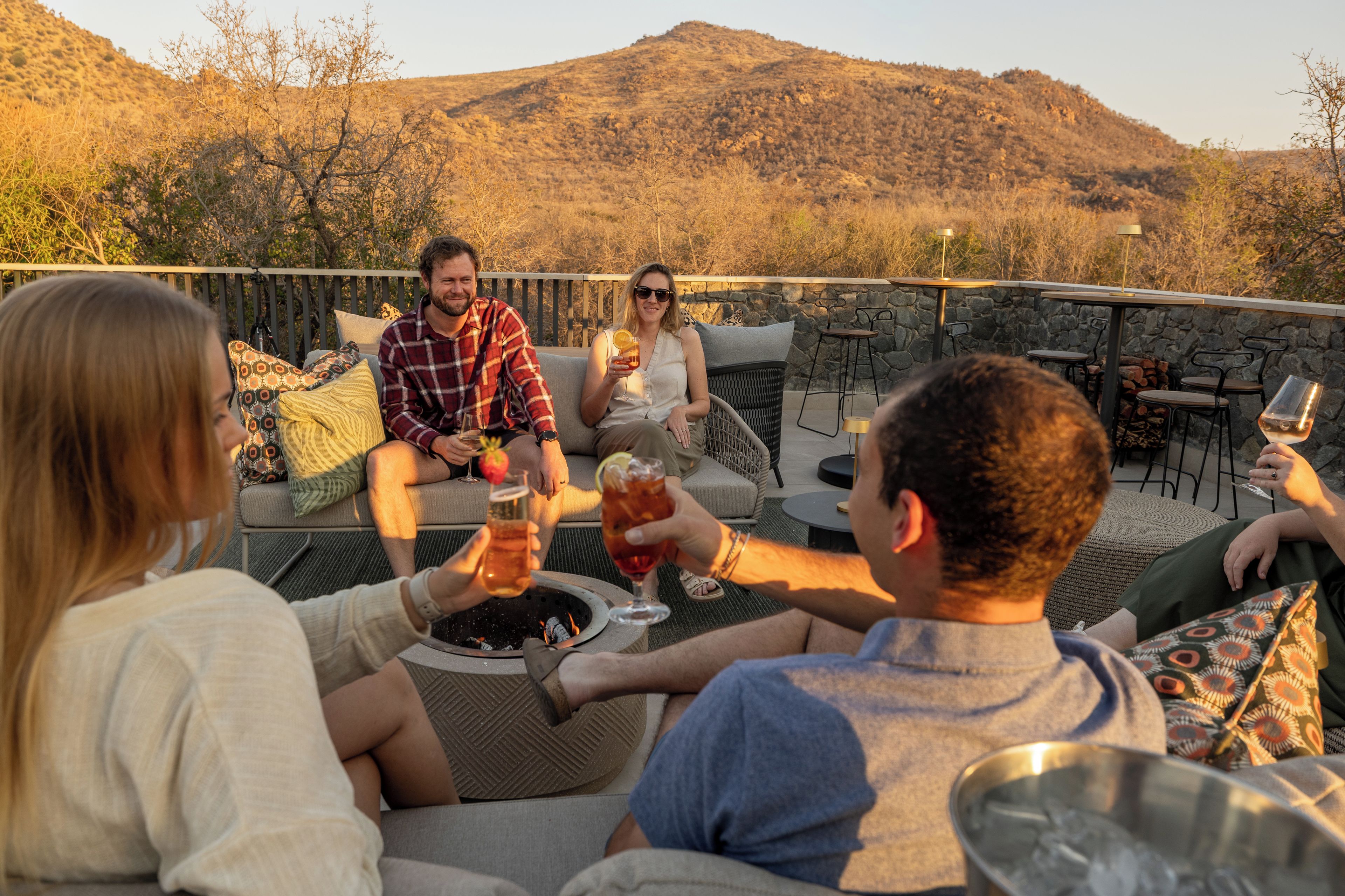 A group of people seated on outdoor furniture around a fire pit, with a mountainous landscape in the background.
