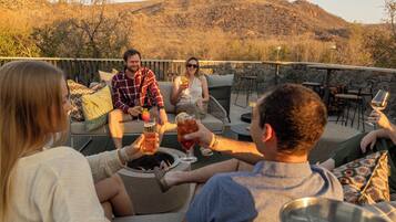 A group of people seated on outdoor furniture around a fire pit, with a mountainous landscape in the background.