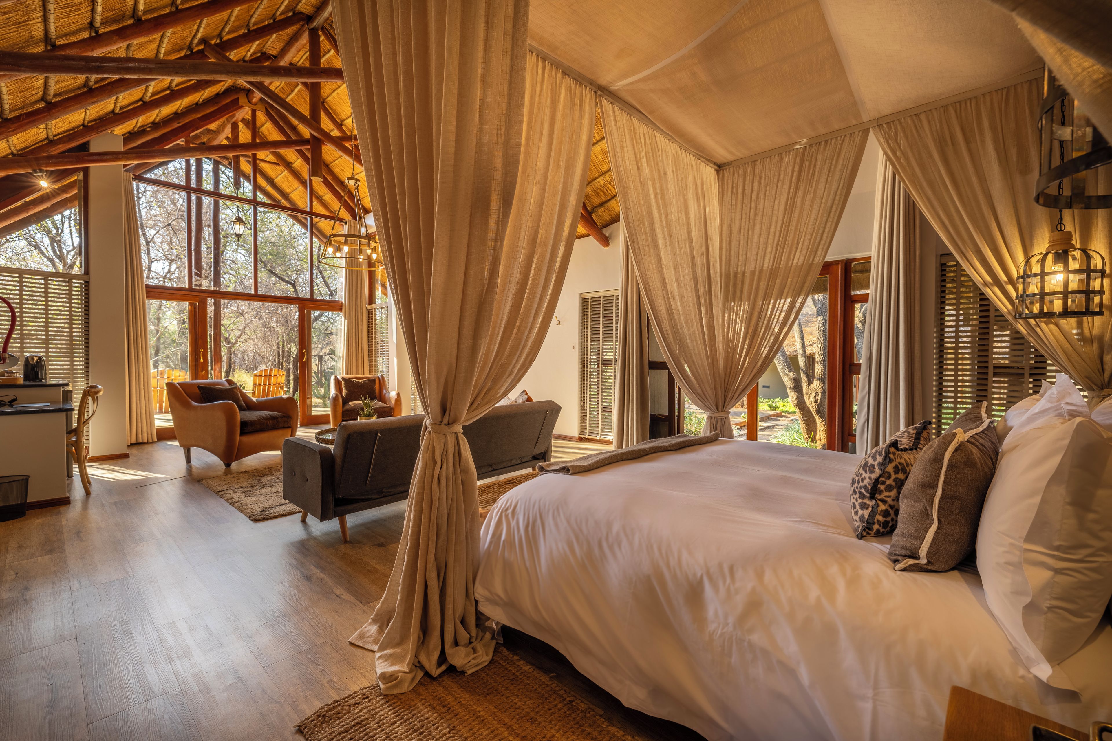 A spacious bedroom with a large bed, a sitting area with a sofa and chairs, and a view of the outdoors through large windows.