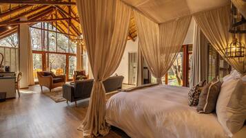 A spacious bedroom with a large bed, a sitting area with a sofa and chairs, and a view of the outdoors through large windows.