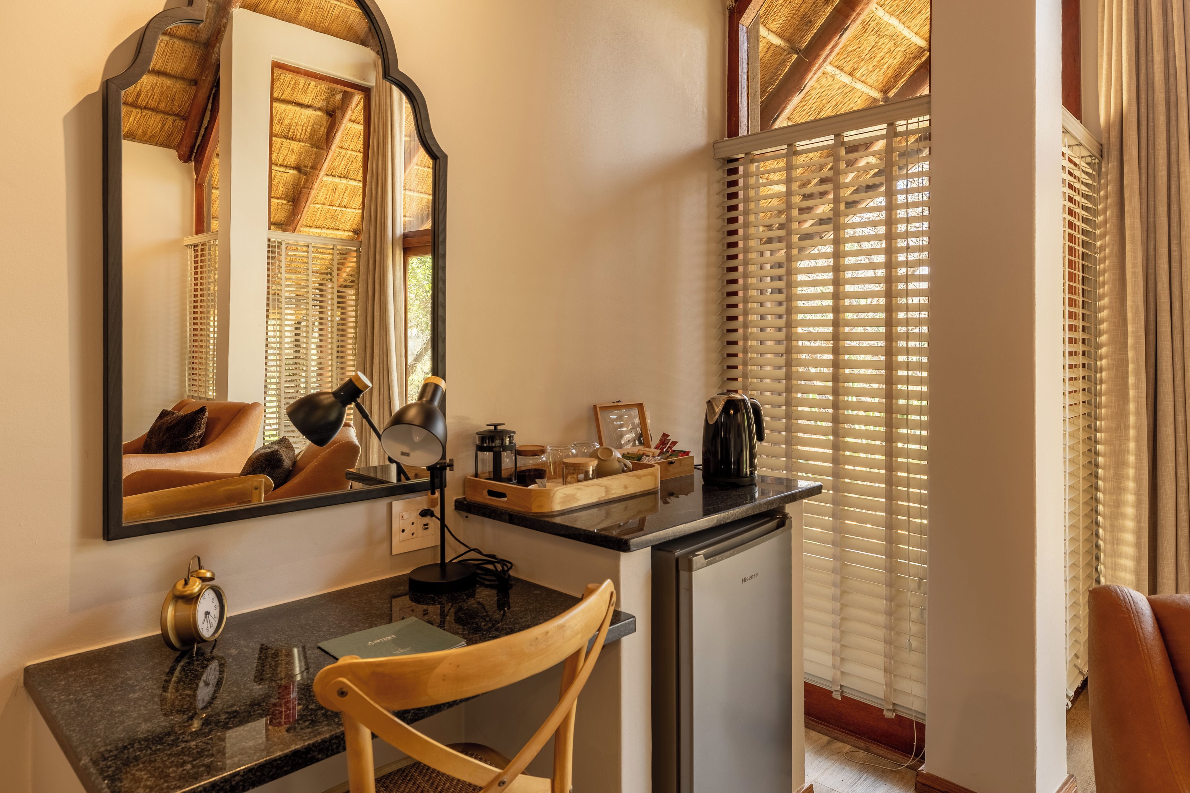 A compact hotel room with a kitchenette, a desk with a chair, a mirror, and a window with blinds.