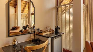 A compact hotel room with a kitchenette, a desk with a chair, a mirror, and a window with blinds.