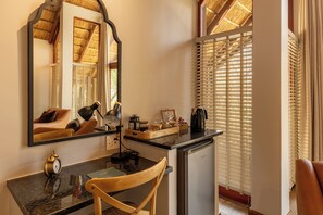 A compact hotel room with a kitchenette, a desk with a chair, a mirror, and a window with blinds. - Tsholo Safari Lodge (Pilanesberg National Park)