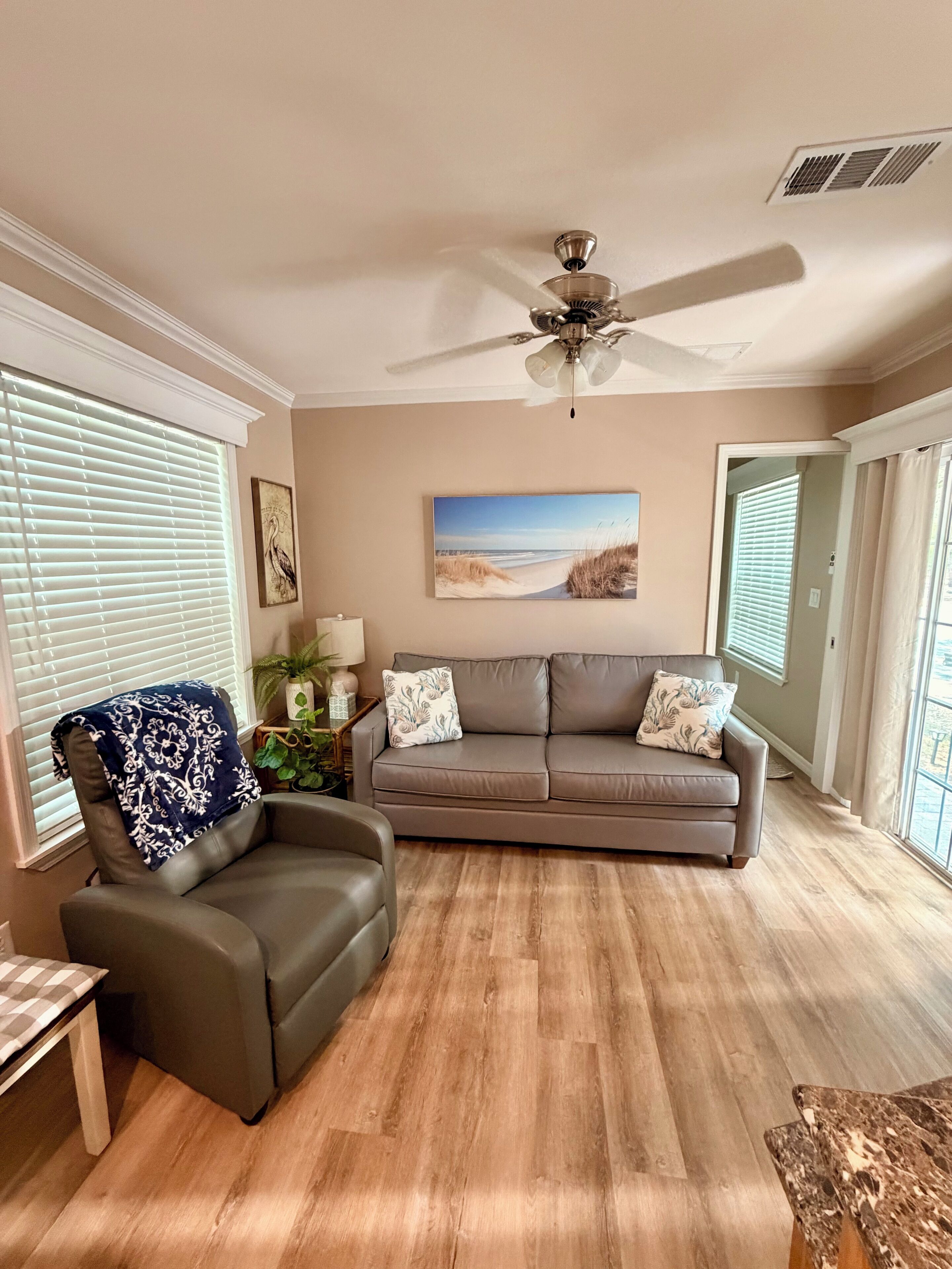 Un salon avec un canapé en cuir marron, un fauteuil à motifs bleus, un ventilateur de plafond et un tableau sur le thème de la plage accroché au mur.