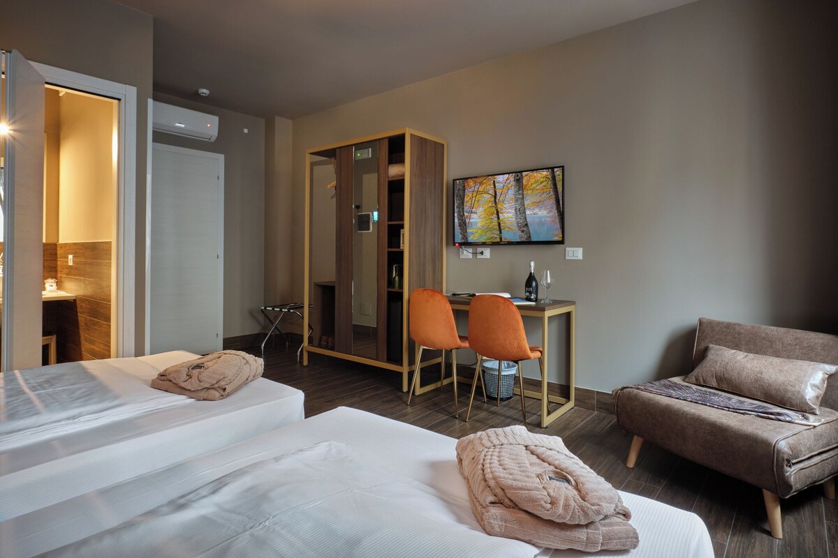 A hotel room with two beds, a desk, a chair, a sofa, and a painting on the wall.