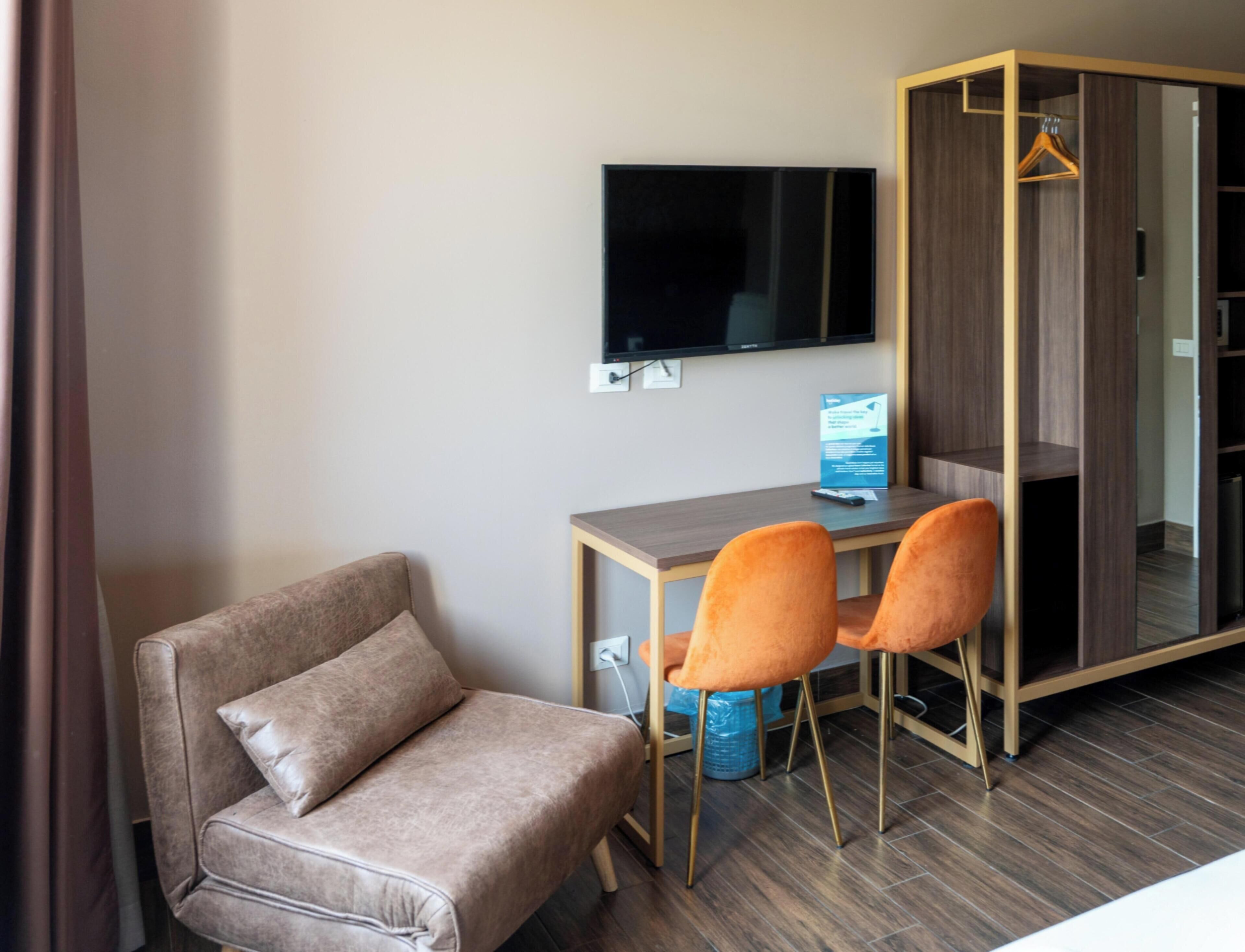 Ein Hotelzimmer mit einer Couch, zwei Sesseln, einem Schreibtisch mit Computer, einem Fernseher und einem Kleiderschrank.